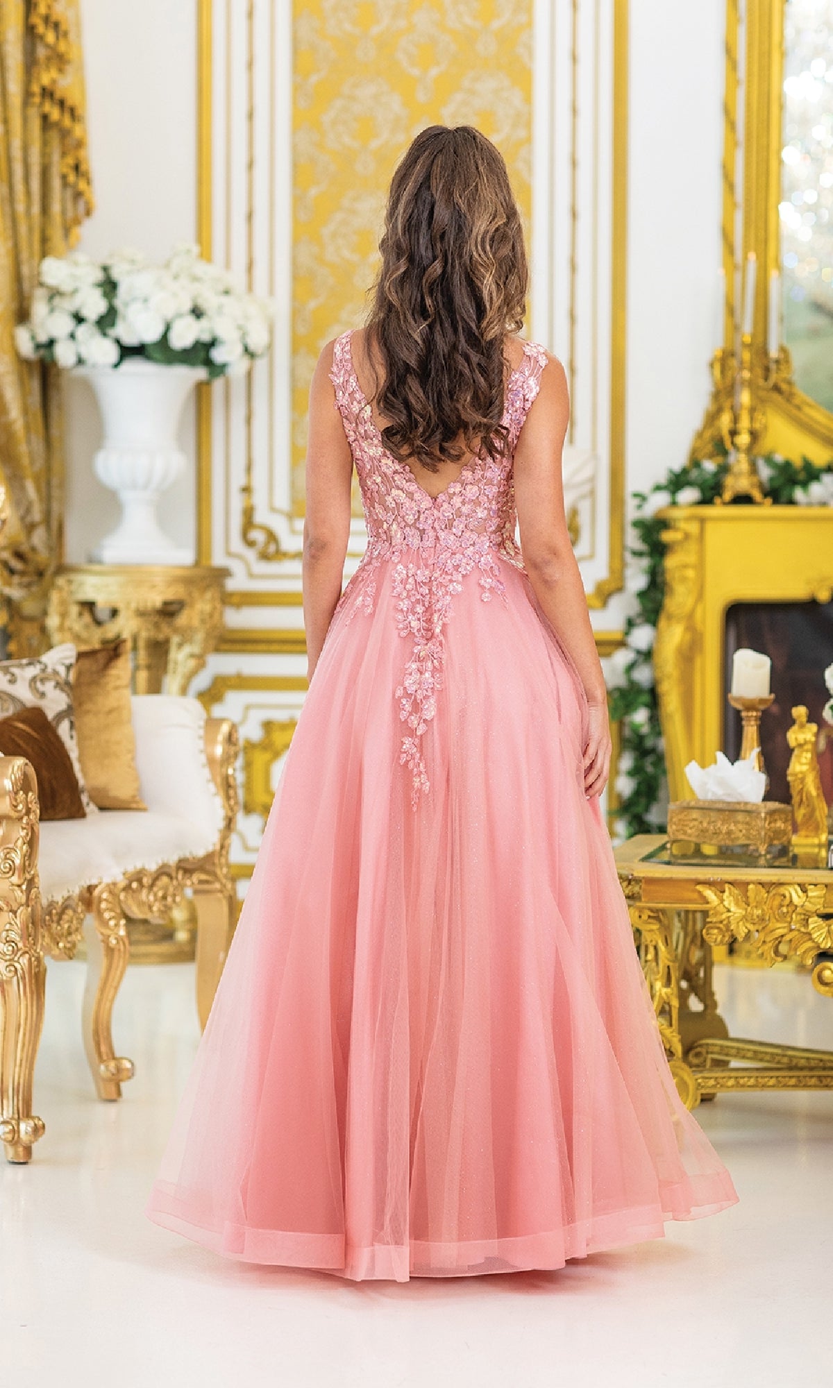 A woman wearing the Dancing Queen 4430 pastel long prom ball gown with sequin embroidery stands in an ornate room with gold accents and white flowers, her back to the camera.